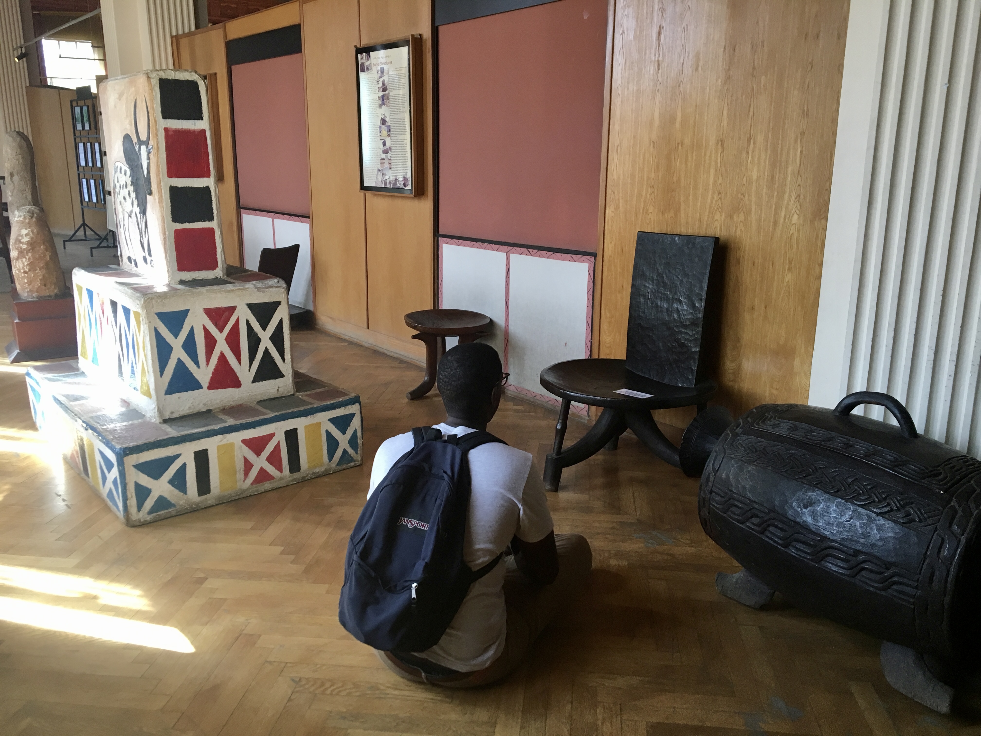 Robell sitting on the floor in a museum in Ethiopia to study a black, locally made chair. He is surrounded by sculptures.
