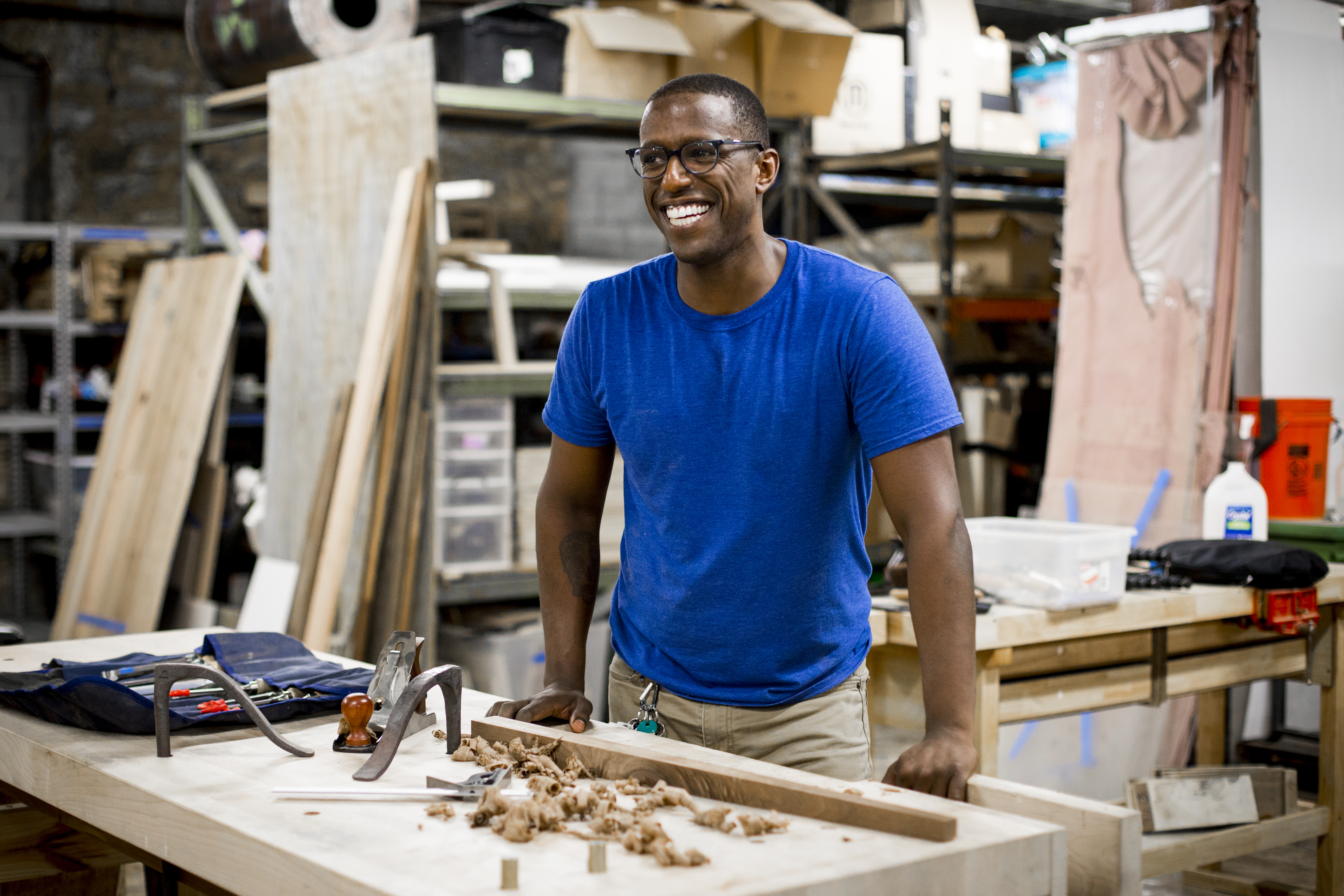 Robell smiling and standing at a workbench. On the bench is a table leg and wood shavings.
