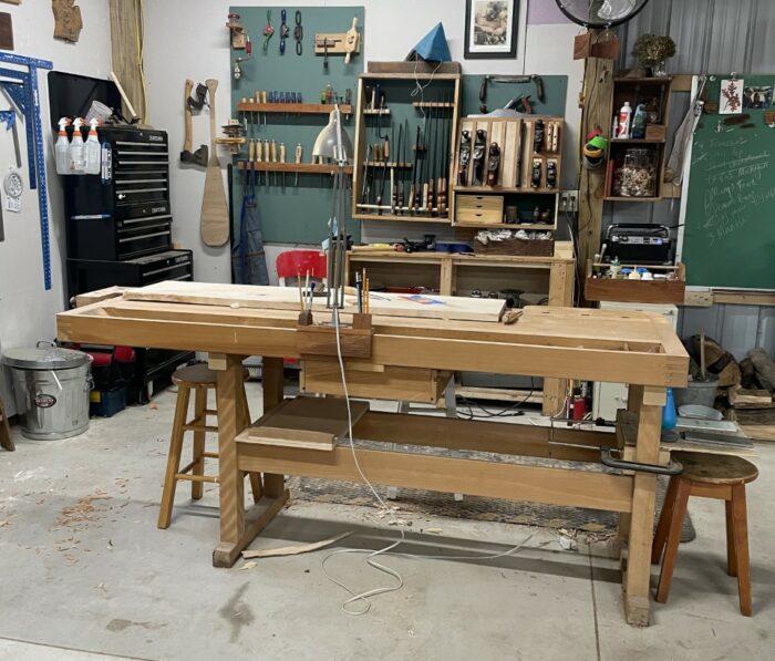 A long workbench in a light-color wood. In the background are green walls and assorted woodworking tools.