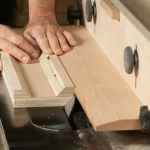 The case pieces should be cut to final width. Use double-sided tape to attach a straightedge perfectly flush with the edge that will be mitered. The L-fence should be set higher than the workpiece and the blade should cut through the workpiece. After cutting the miters, Van Dyke leaves the straightedges taped on.