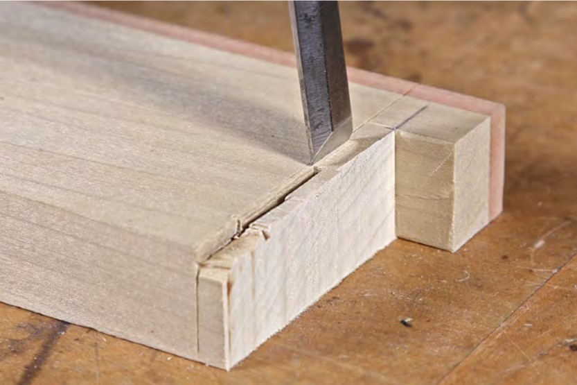 A chisel chopping down into the end of the end of a narrow piece of light-colored wood.