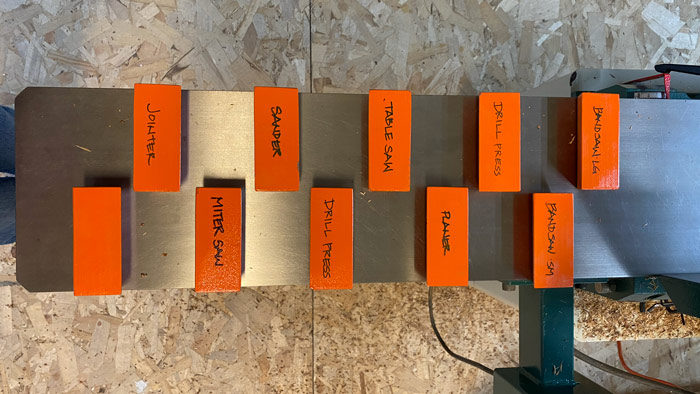 Orange blocks lined up on top of a jointer bed. Each one has a machine or funny saying written on them.