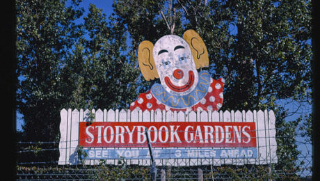 A hand drawn sign with a clown on it above "Storybook Gardens" emblazoned on a white picket fence.