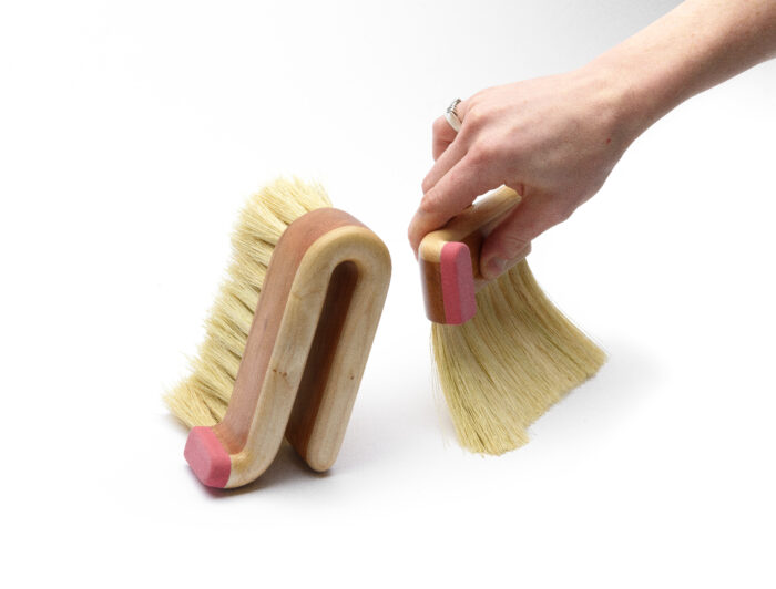 two curved brushes made of maple and cherry wood. A hand is sweeping with one brush.