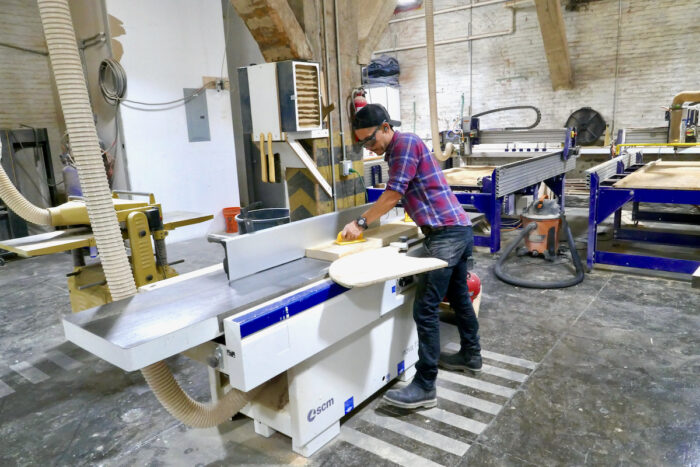 The Ottra Gallery wood shop, including a wide jointer and planer, a panel table saw, shapers, a long bar clamp dock, and a vast CNC machine. An employee uses the jointer.
