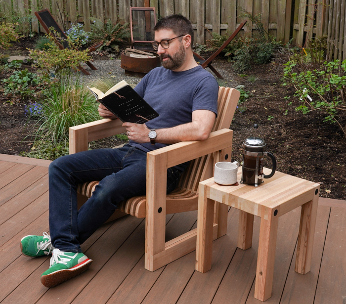 A young white male sits in a chair built to match this outdoor table. Next to him is the table with a coffee cup and a french press. 