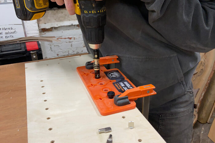 Larissa is drilling a line of perfectly straight holes using a drill and a jig with a line of holes that registers the drill exactly where in needs to be for a straight line of holes. The color of the jig is that of an over-ripe orange.