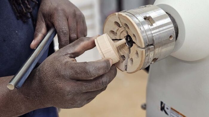 Darryl inserts a top into a new chuck. This chuck is a beefy mechanical four-jaw chuck that looks like it will eat the piece of wood, but the hungry metal jaws have been replaced by much softer looking wood jaws.
