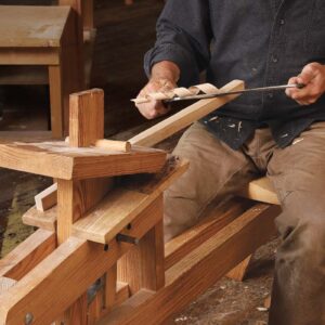 A piece of wood is held in a shaving horse. A woodwoker using a drawknife, a long blade suspended between to handles to pull a perfect curl of wood.