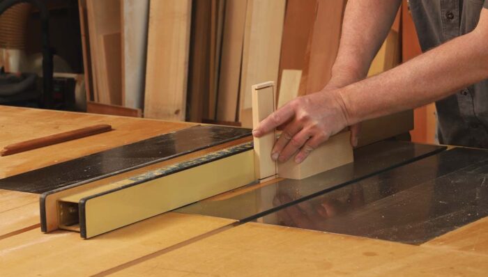Rabbeting cuts are being done at the table saw. A board is being held on end and a shallow cut is taken. The author follows behind the board with larger piece of wood to hold the piece against and steady.