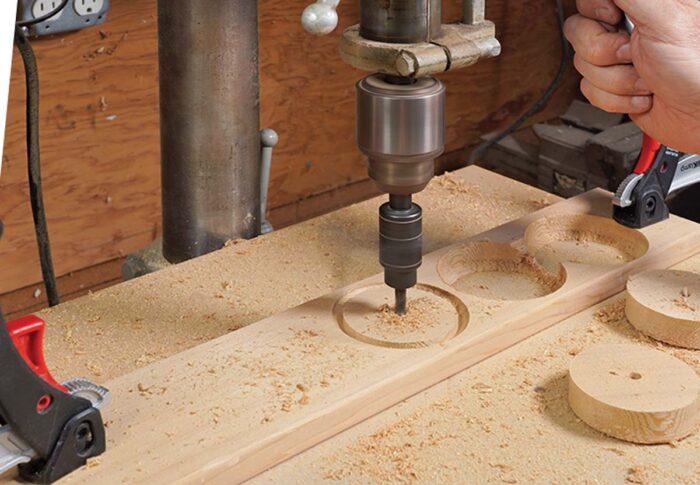 Asa cuts circles on the drill press with the circle cutting jig. a couple pieces are already set aside, and the work piece is clamped to the table. 