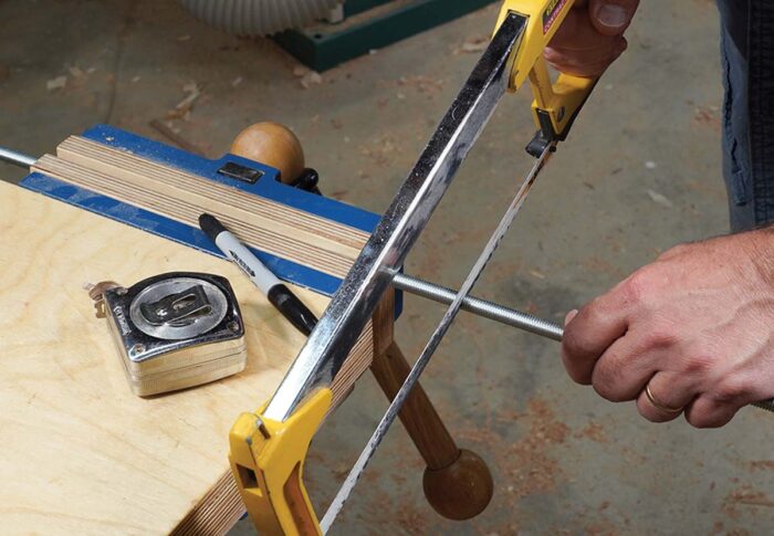 Asa uses a hacksaw to cut threaded rod to length. The rod is held in a vise to make cutting easier.