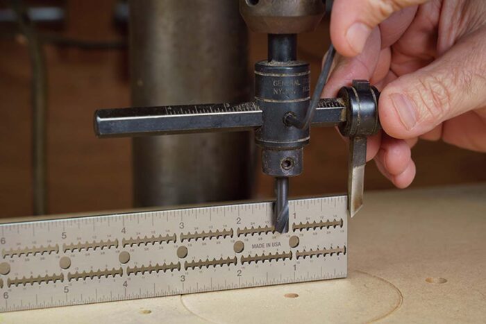 Asa holds a circle cutting jig made for the drill press. The cutting arm is adjustable, and he holds the edge over a ruler to get the correct radius.