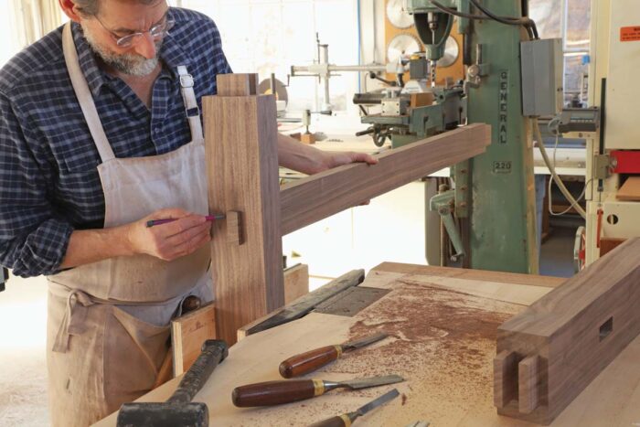 One of the legs sits in a vice while tom holds the stretcher in place. He marks the depth of the proud tenon, to indicate where he should profile the tenon edge.
