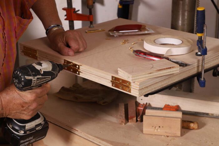 Beth uses a drill to secure the hinge to the edge of two pieces of plywood.