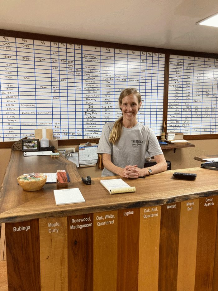 Bonnie stands behind a counter in a lumberyard office. Samples of different wood species cover the front of the counter, and 