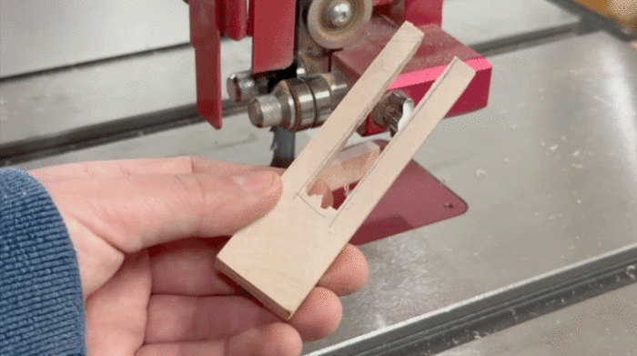 Amanda holds the small piece of wood in front of the bandsaw, and has cutaway the center of the board. The two remaining prongs stand straight, indicating the board is stable.