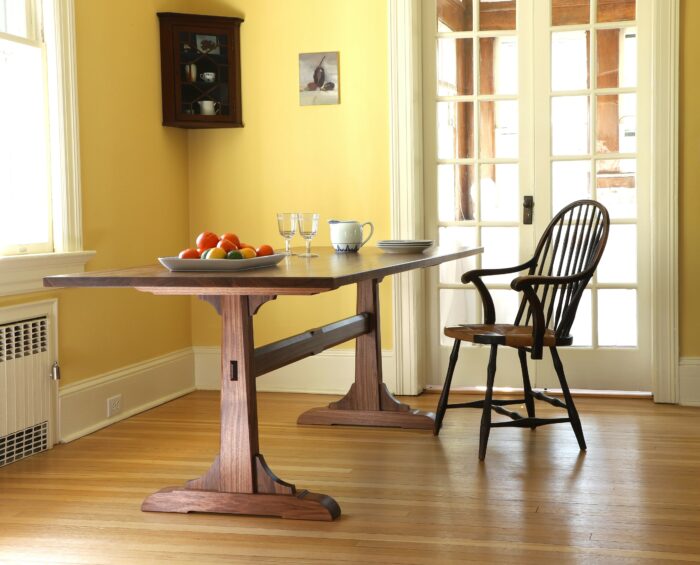 The walnut trestle table sits in a dining room with dishes on the table and a Windsor chair nearby.