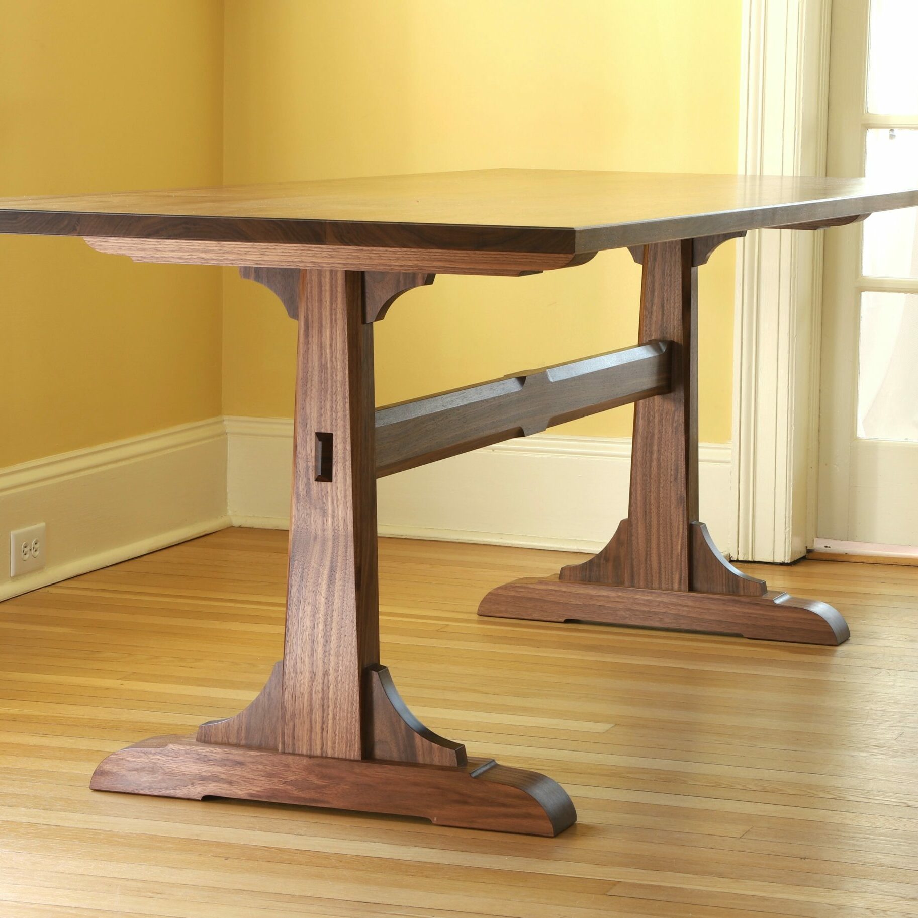 A walnut trestle table sits in a room with yellow walls and a hardwood floor.