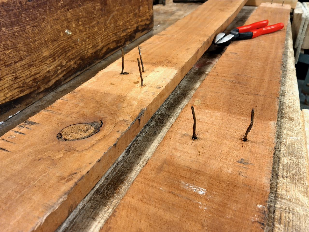 Nails sticking straight up through a board, with pliers sitting in the background.