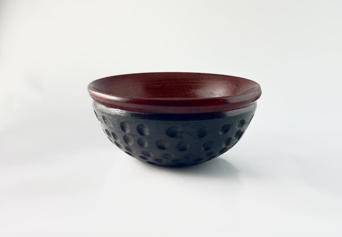 A padouk bowl with a rounded edge at the top and divots carved into the sides of the bowl. The rim of the bowl is left natural and the rest of the bowl is dyed black.