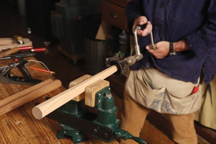 A chair leg is held in a vise as a cone-shaped tool is spun around the end with a brace. 