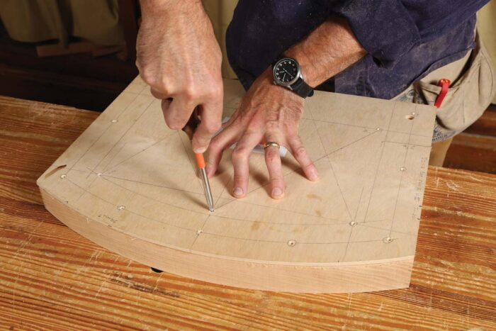 A wooden template is on the seat blank and an awl is being used to transfer holes from the template onto the seat blank. 